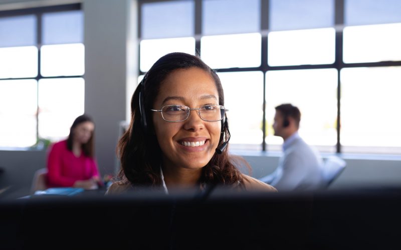 Front view close up of a mixed race woman working as a customer support, wearing smart clothes and headset, using a desktop computer with colleagues working in the background.